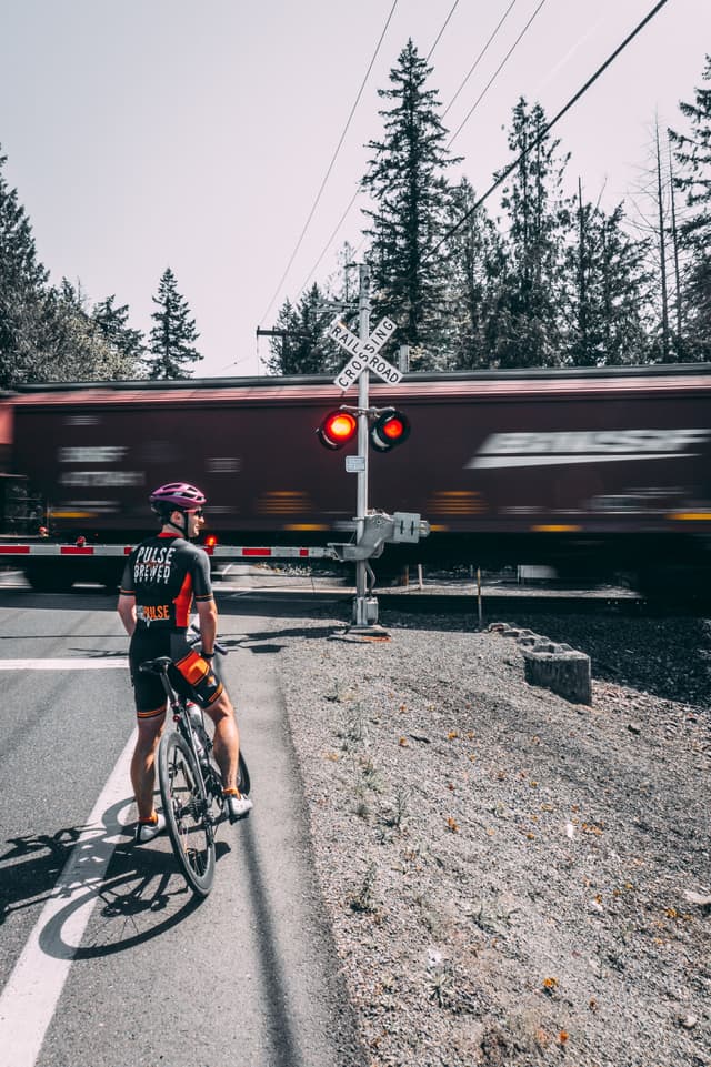 A cyclist waits at a railroad crossing as a train passes by, with red lights flashing and surrounded by trees