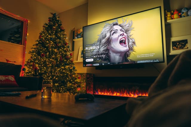 Living room with Christmas tree and lit fireplace, TV displaying a dramatic scene