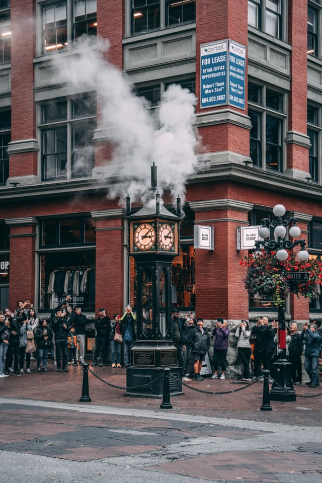 A street scene featuring a crowd of people gathered around a steam clock, with smoke billowing from its top. The clock is set against an urban backdrop of a red-brick building, adorned with hanging flower baskets and street lamps