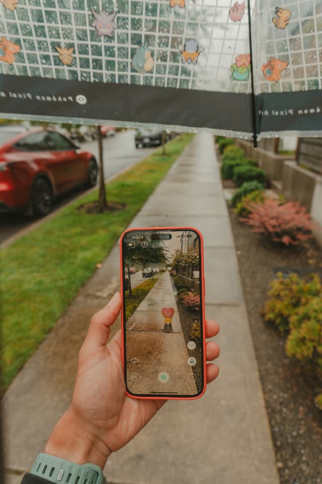 A person holding a smartphone under an umbrella, capturing a photo of a rainy sidewalk with augmented reality elements on the screen