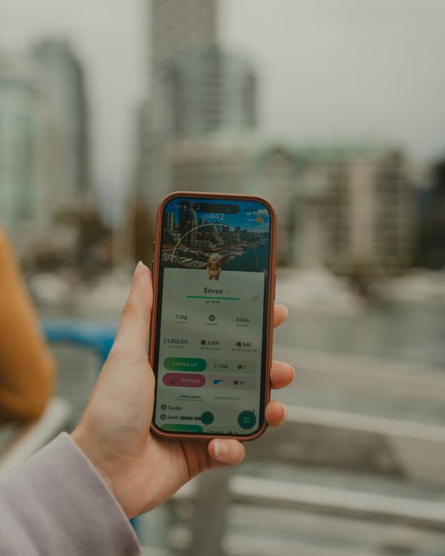 A hand holding a smartphone displaying an augmented reality game, with a blurred cityscape in the background