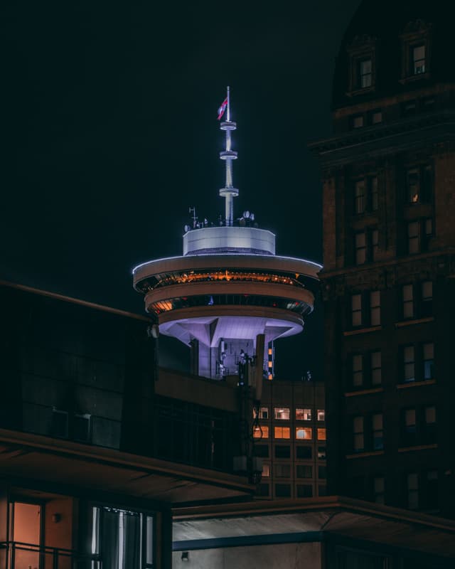 A tall, illuminated tower with a circular observation deck and spire, surrounded by dark buildings in a nighttime urban setting
