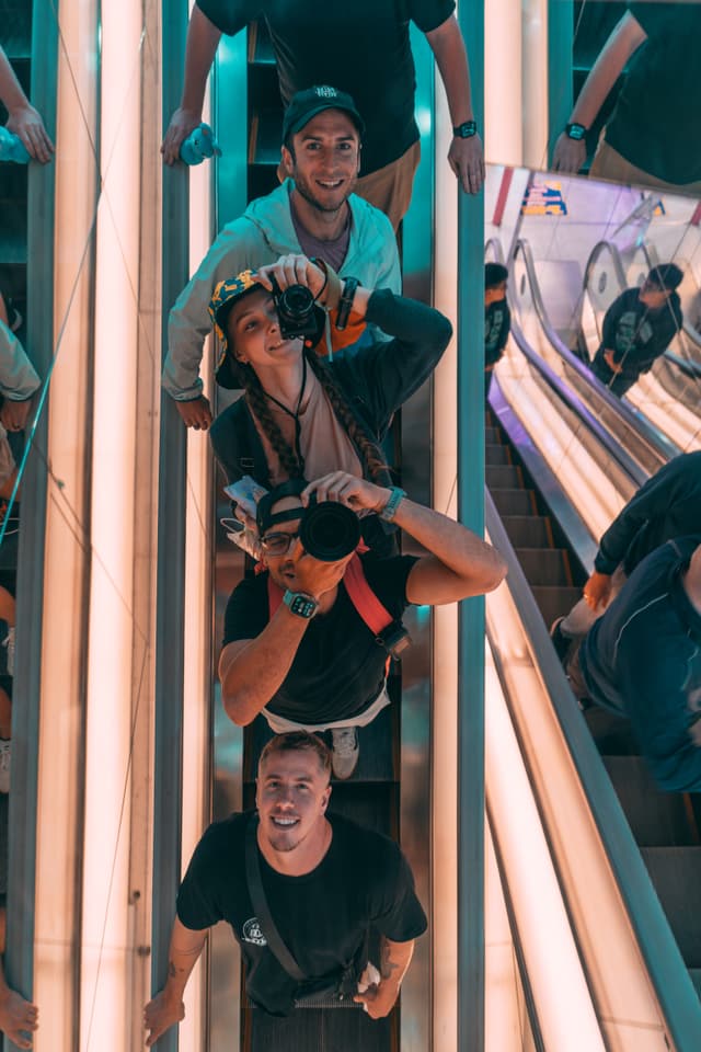 A group of people on an escalator, with some holding cameras and looking up, surrounded by mirrored surfaces
