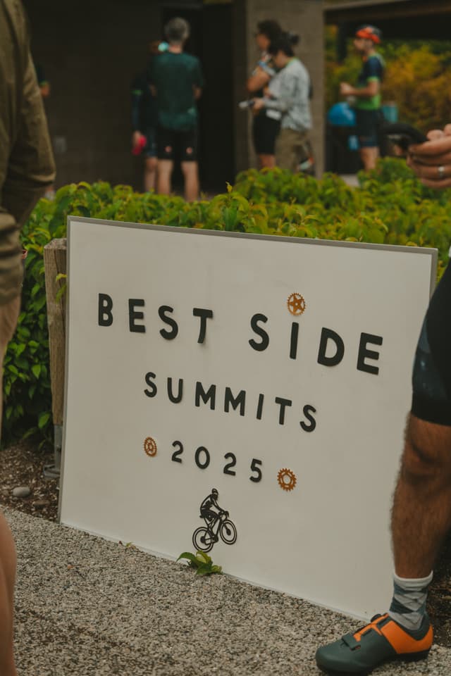 A sign reading BEST SIDE SUMMITS 2025 with a bicycle graphic, surrounded by people and greenery in an outdoor setting