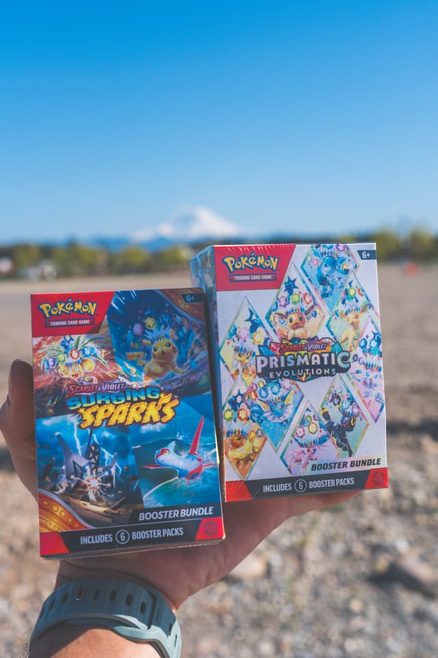 A hand holding two Pokémon booster bundle boxes, Surging Sparks and Prismatic Evolutions, with a scenic outdoor background