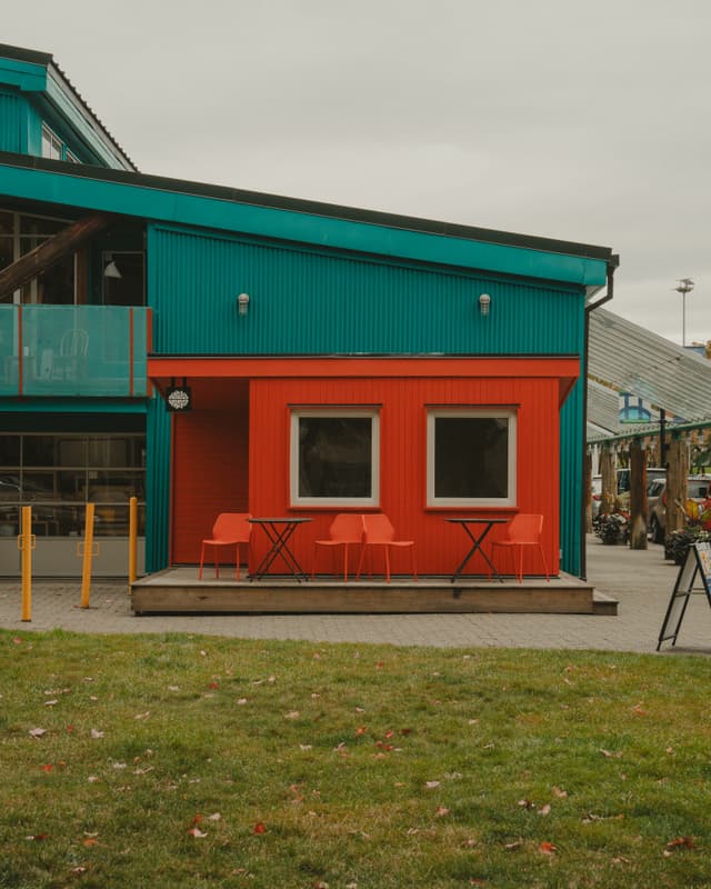 A vibrant building with turquoise and red exterior features, including two windows and orange chairs at small tables. The foreground has a grassy area, and the sky is overcast