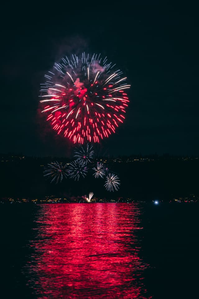 Fireworks exploding in the night sky over a body of water, with reflections of red and white lights on the surface