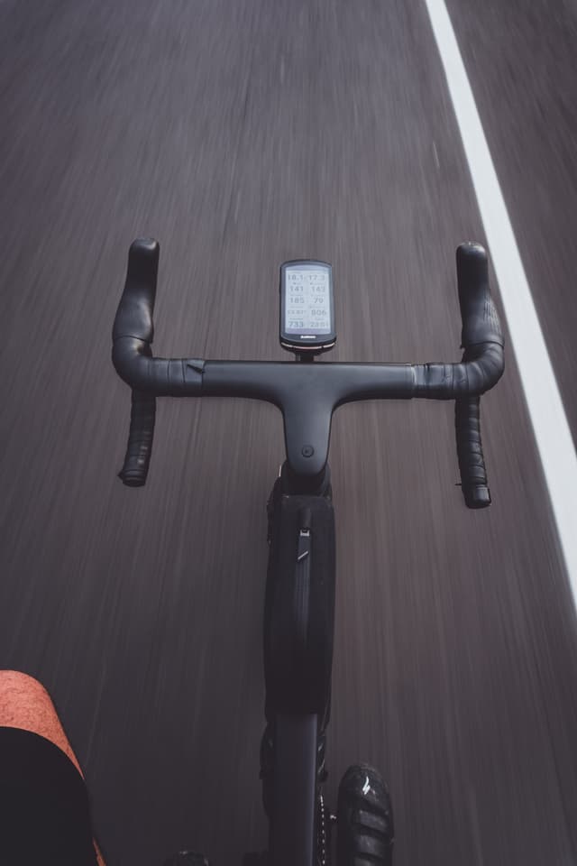 A cyclist's view of a road, featuring bicycle handlebars with a mounted device, and a white line marking on the pavement