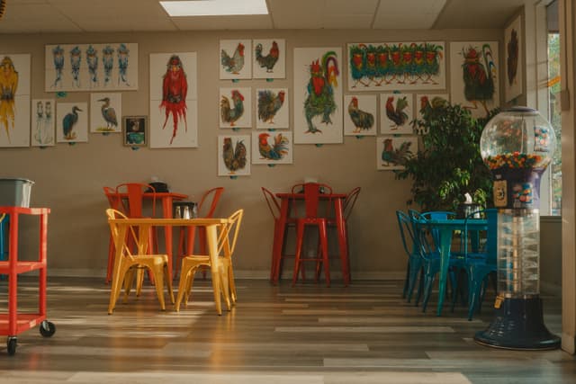 A colorful, eclectic cafe interior with mismatched chairs and tables, vibrant wall art featuring roosters, and a gumball machine in the corner