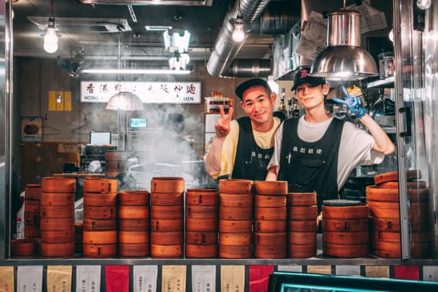 Two people in a food stall with stacks of bamboo steamers, smiling and posing for the camera