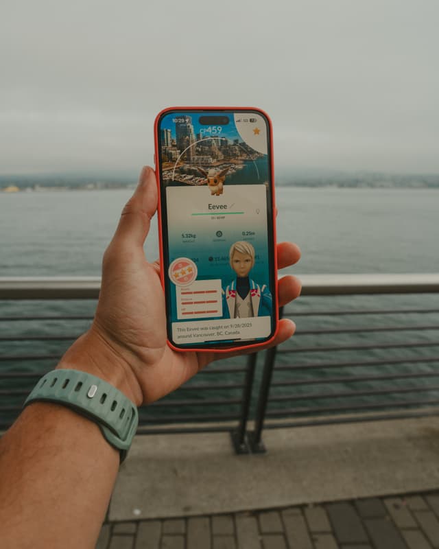 Hand holding a smartphone displaying a Pokémon screen, with waterfront railing and cloudy sky background