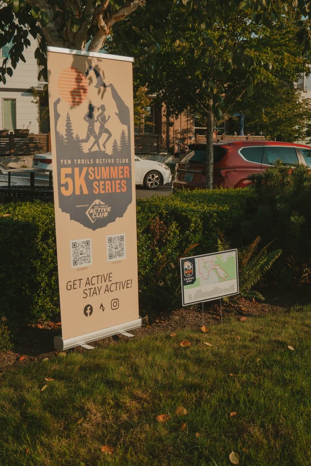 A banner advertising a 5K summer series event with the tagline Get Active, Stay Active stands on a grassy area next to a smaller sign with a map Cars and trees are in the background