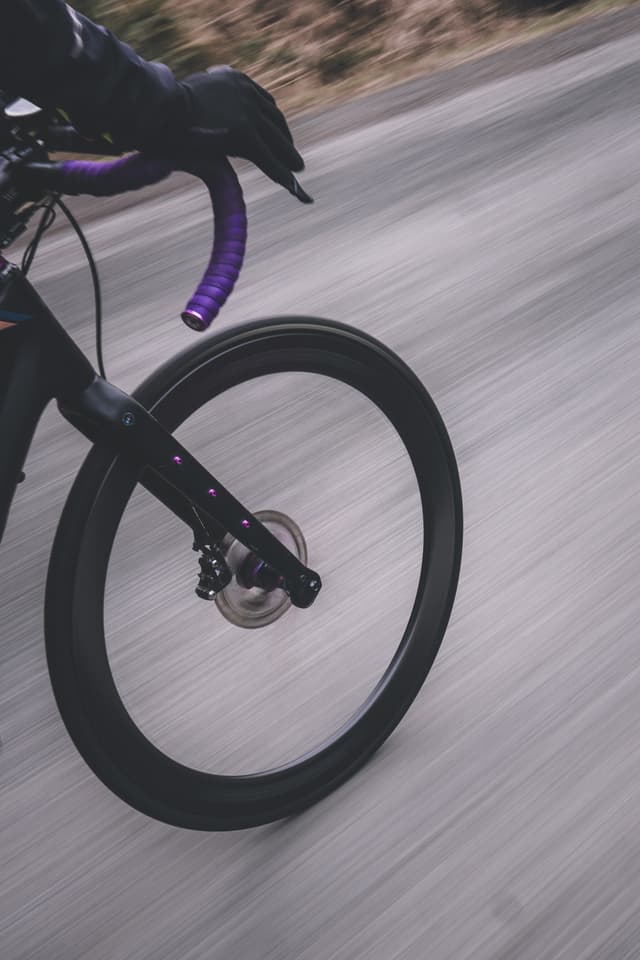 A close-up of a bicycle wheel in motion on a road, with a gloved hand on the handlebars