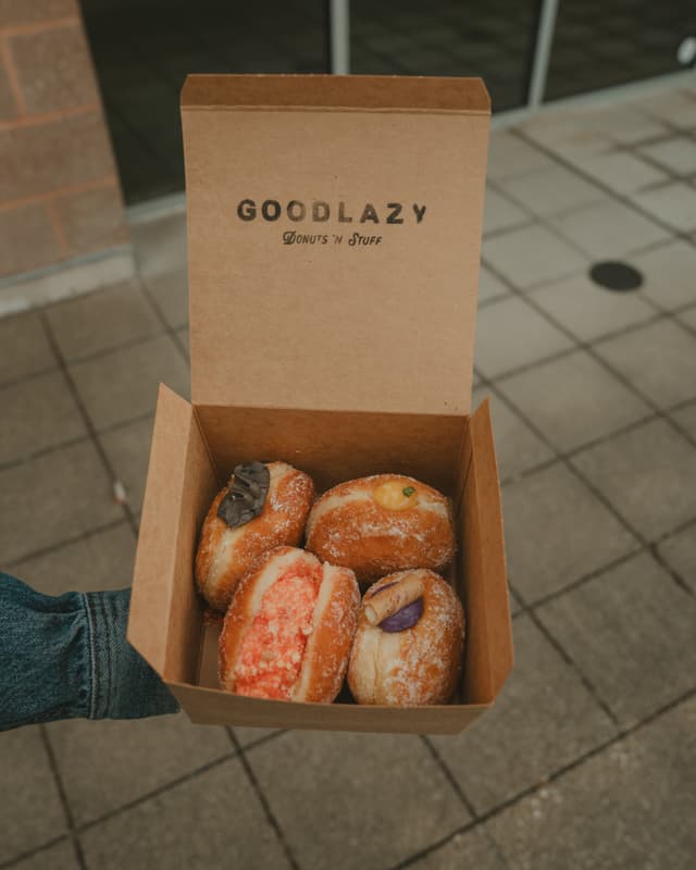 A hand holding an open box containing four assorted filled doughnuts, with GOODLAZY printed on the inside lid