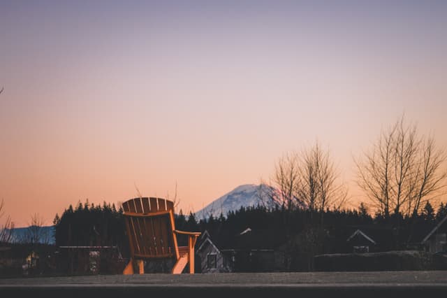 A wooden chair facing a snow-capped mountain at sunset, with bare trees and rooftops in the foreground