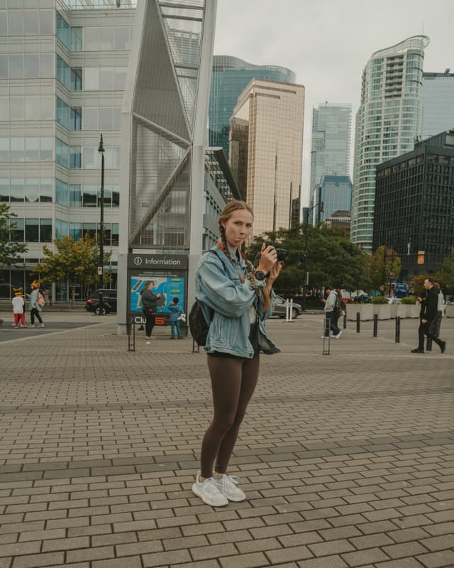 An individual with a camera stands in a city plaza surrounded by modern architecture and skyscrapers