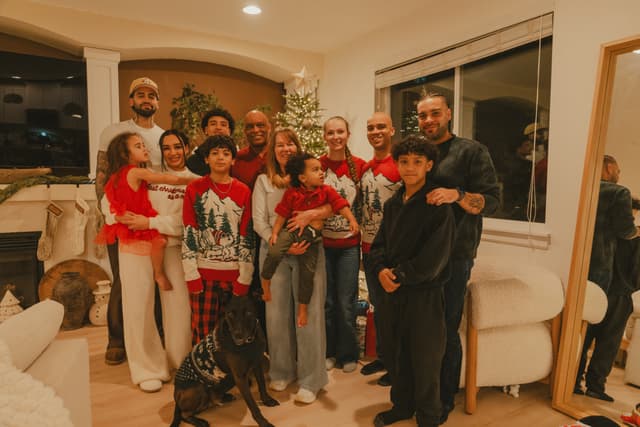 Large family gathered in a warmly lit living room wearing festive Christmas pajamas and outfits, posing together with a dog in front of a decorated tree and fireplace