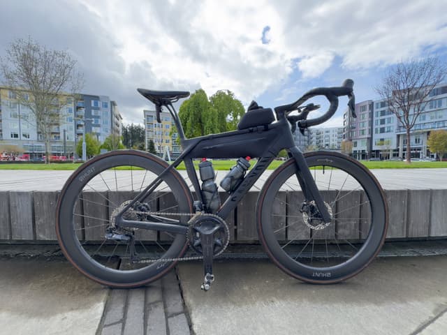 A sleek black canyon endurace road bike is parked on a concrete surface with modern buildings and trees in the background under a cloudy sky