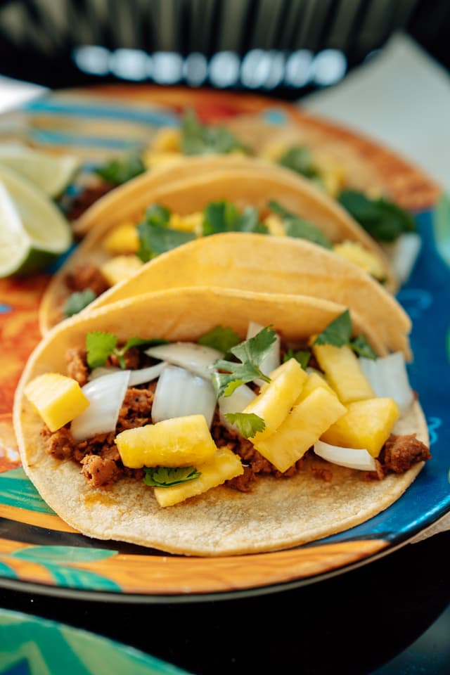 Three tacos filled with meat, pineapple, onions, and cilantro on a colorful plate, with lime wedges in the background