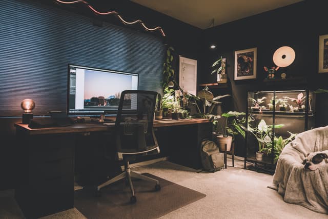 A cozy home office with a large desk, computer, ergonomic chair, and various plants The room has dark walls, soft lighting, and decorative elements like framed pictures and a doughnut light