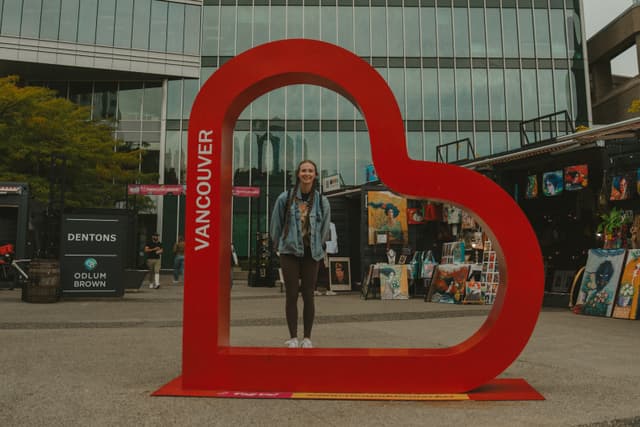Person standing inside a large red heart-shaped frame in an urban setting with buildings and market stalls in the background