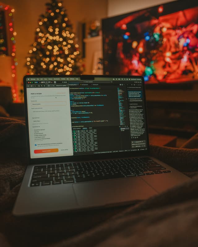 Open laptop displaying code in a dimly lit living room with a glowing Christmas tree and colorful TV screen in the background, creating a cozy holiday work atmosphere