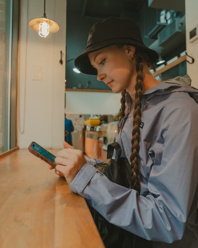 A person with braided hair wearing a bucket hat and jacket is sitting at a wooden counter, looking at a smartphone. A warm light hangs above, and there are shelves and windows in the background