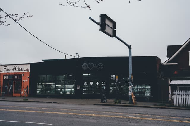 A street scene featuring a row of buildings, including a black storefront with large windows and a red building to the left A traffic light and power lines are visible above the street