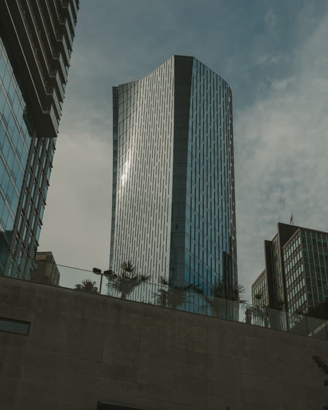 A modern skyscraper with reflective glass facade against a cloudy sky, surrounded by smaller buildings