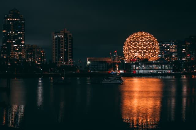 A nighttime cityscape featuring illuminated buildings along a waterfront A prominent geodesic dome glows brightly, reflecting off the calm water