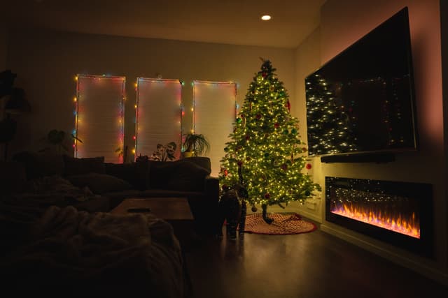 A cozy living room decorated for Christmas, featuring a lit Christmas tree, a fireplace, and string lights on the windows