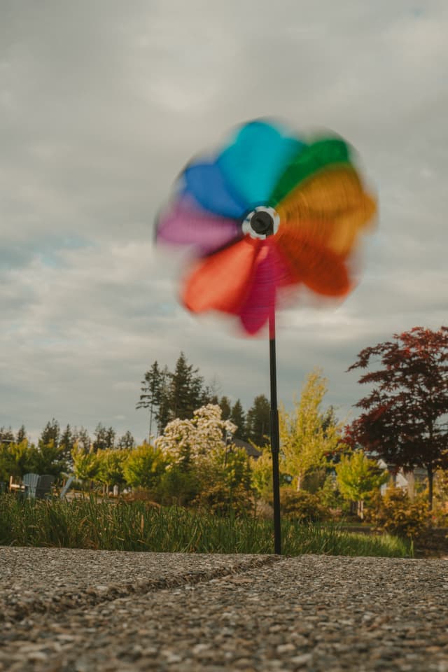 A colorful pinwheel spinning in a garden setting with trees and a cloudy sky in the background
