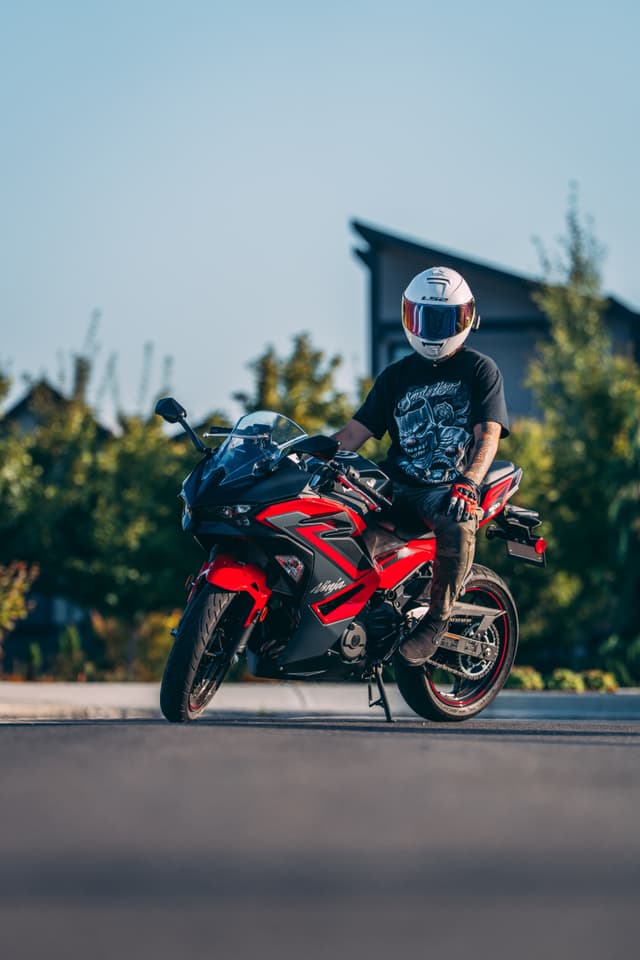 A person wearing a helmet and dark clothing sits on a red and black sport motorcycle on a paved surface with greenery in the background
