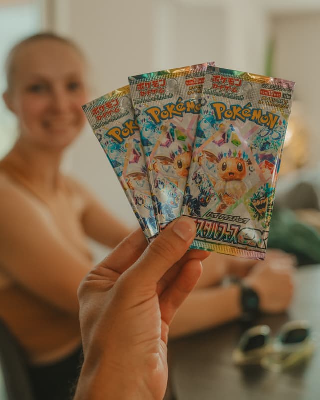 A hand holding three Pokémon card packs with a smiling person in the background