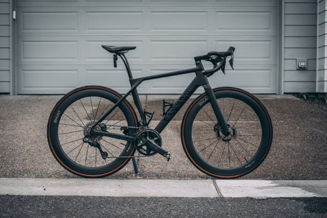 A sleek black road bike with drop handlebars and disc brakes, positioned in front of a closed garage door on a concrete driveway