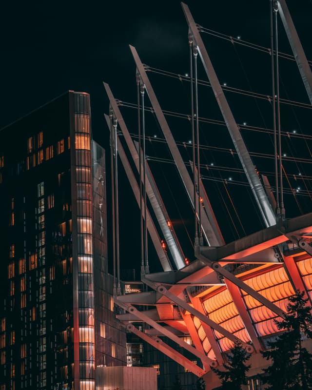 A modern architectural structure with angled beams and illuminated orange panels, set against a dark sky A nearby building with lit windows is visible