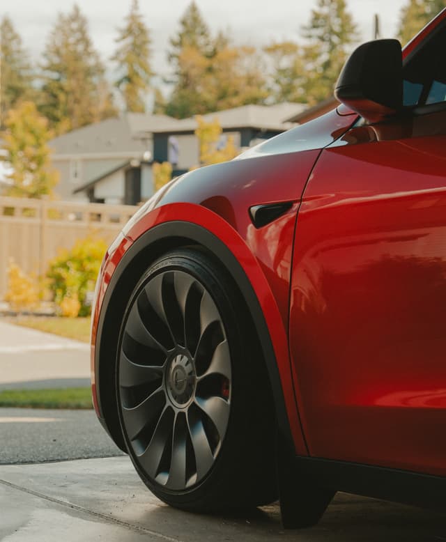 A red car with sleek, modern wheels is parked in a driveway, with houses and trees visible in the background