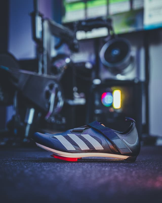 A cycling shoe is placed on a floor with an indoor cycling bike and gym equipment in the background