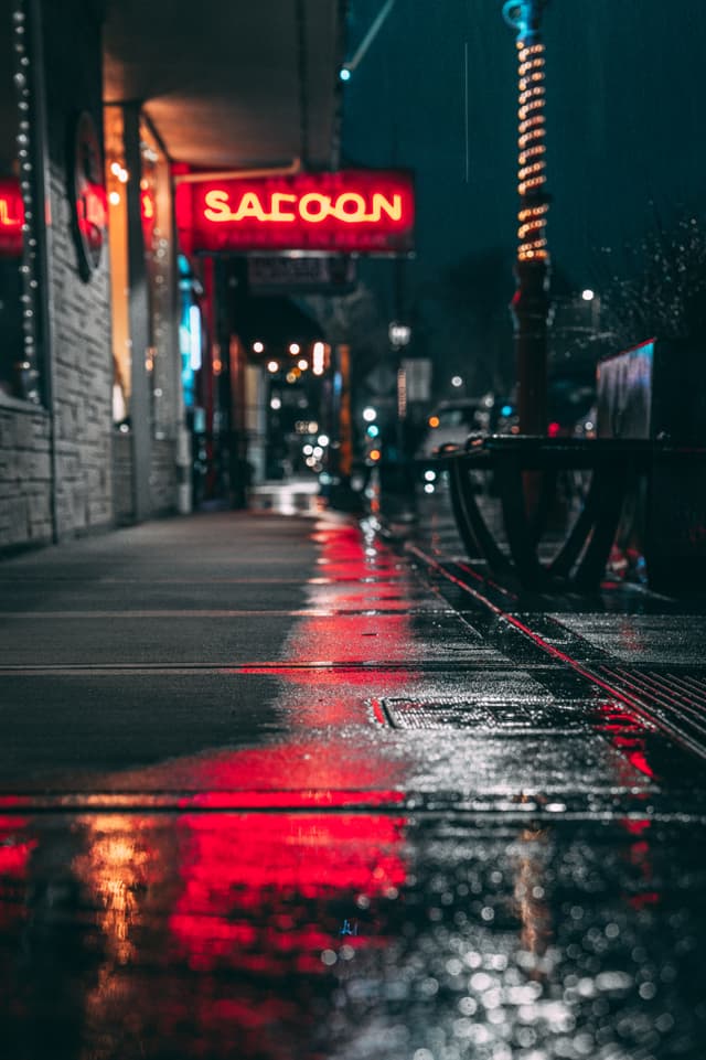 A wet city sidewalk at night with reflections of neon lights, including a red SALOON sign, creating a moody urban atmosphere