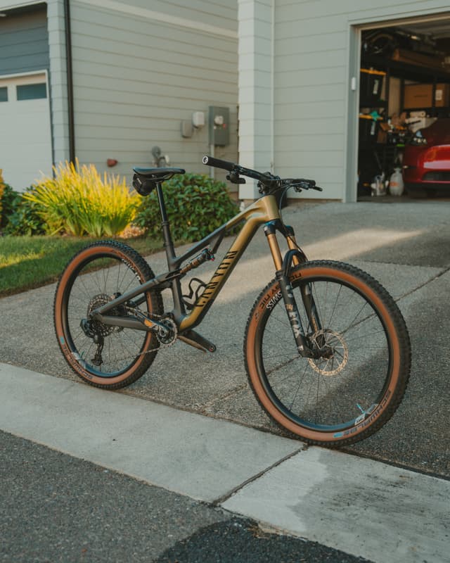 A mountain bike with a gold and black frame is parked on a driveway in front of a garage