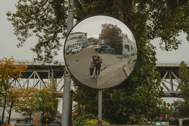 A convex mirror reflects two people on a pathway with trees and an industrial structure in the background