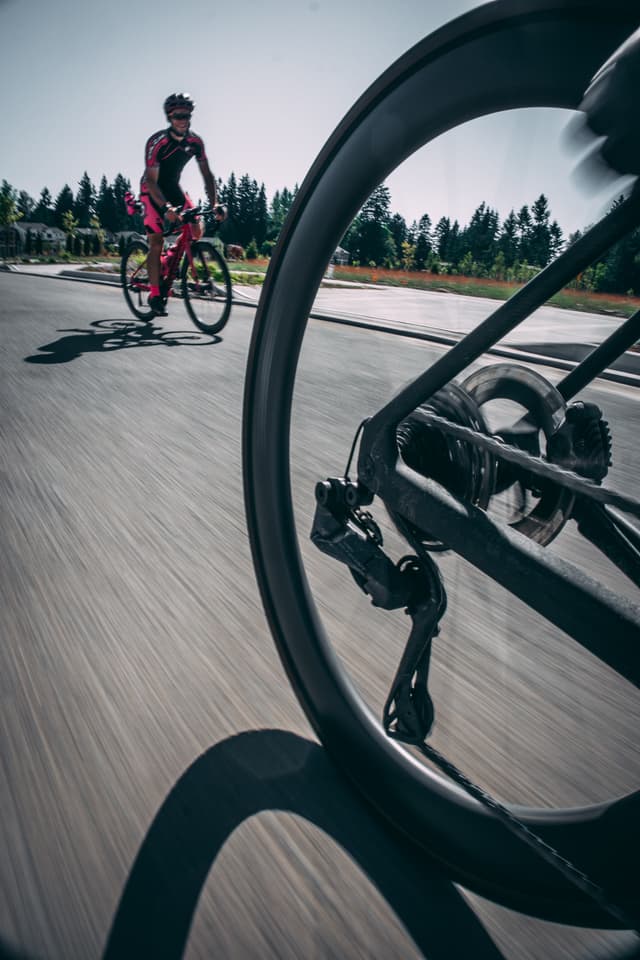 A cyclist in pink rides on a road, viewed through the spokes of another bicycle's wheel in the foreground