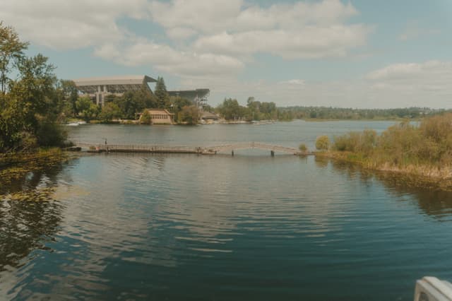 A serene lake with a stadium in the background, surrounded by trees and vegetation under a partly cloudy sky