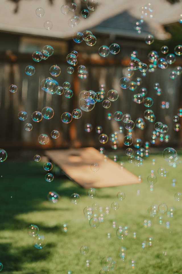 Numerous bubbles floating in a backyard with a wooden fence and a cornhole board in the background