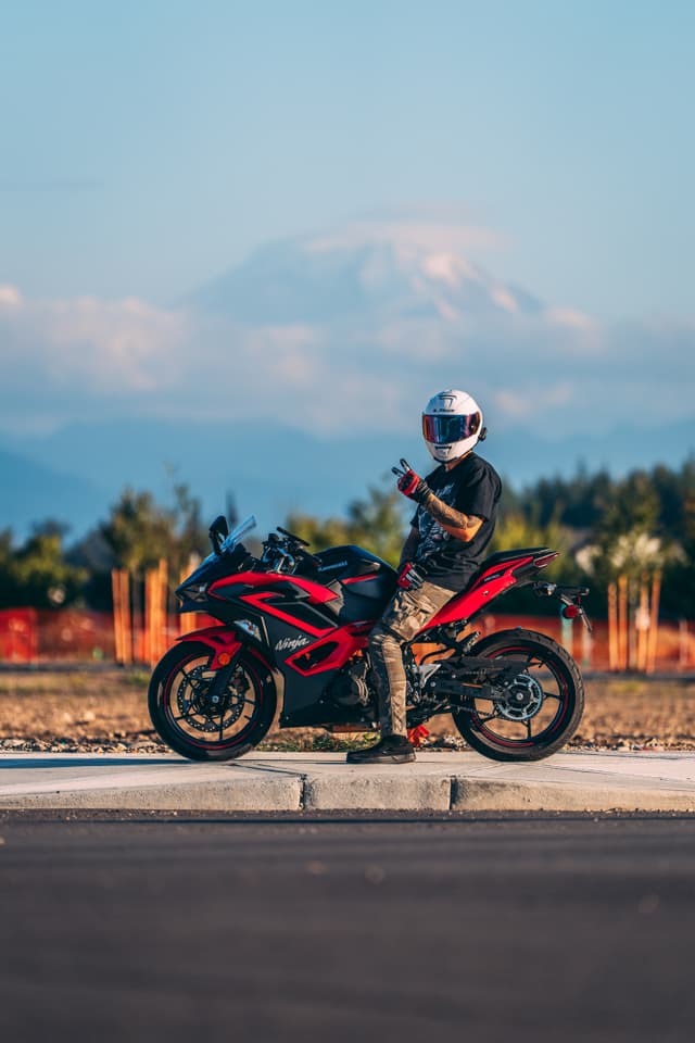 A person wearing a helmet sits on a red sport motorcycle on the side of a road, with mountains and trees in the background