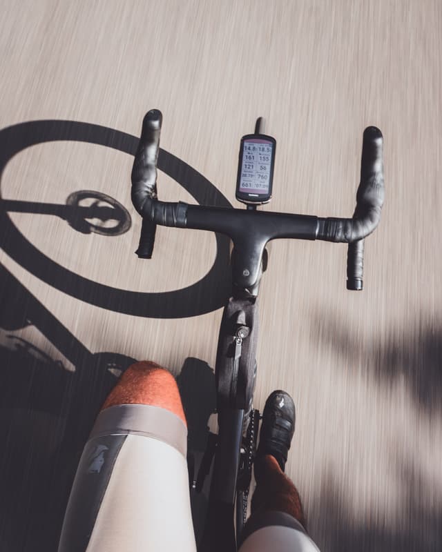 A cyclist's perspective from above, showing the handlebars with a mounted device, legs in motion, and the shadow of the bicycle on the road