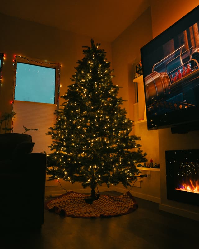 A lit Christmas tree by a fireplace and a large TV in a cozy living room