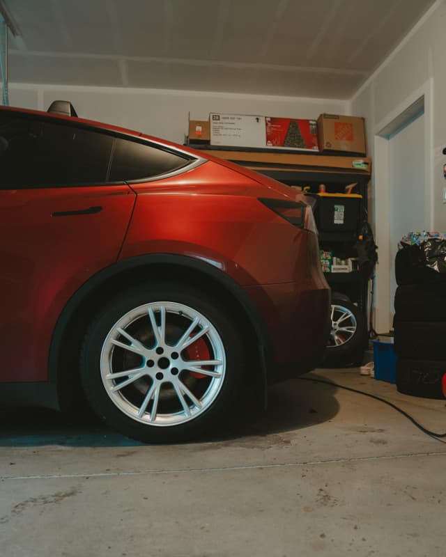 red Tesla parked in a home garage plugged into a charger with shelves and tools visible in the background