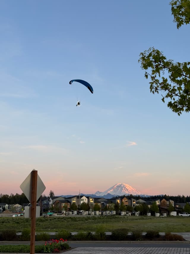 A paraglider is soaring in the sky above a suburban area with a snow-capped mountain in the background during sunset