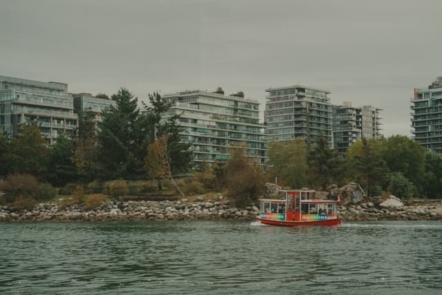 A small red and yellow boat is on the water near a rocky shoreline with apartment buildings and trees in the background. The sky is overcast